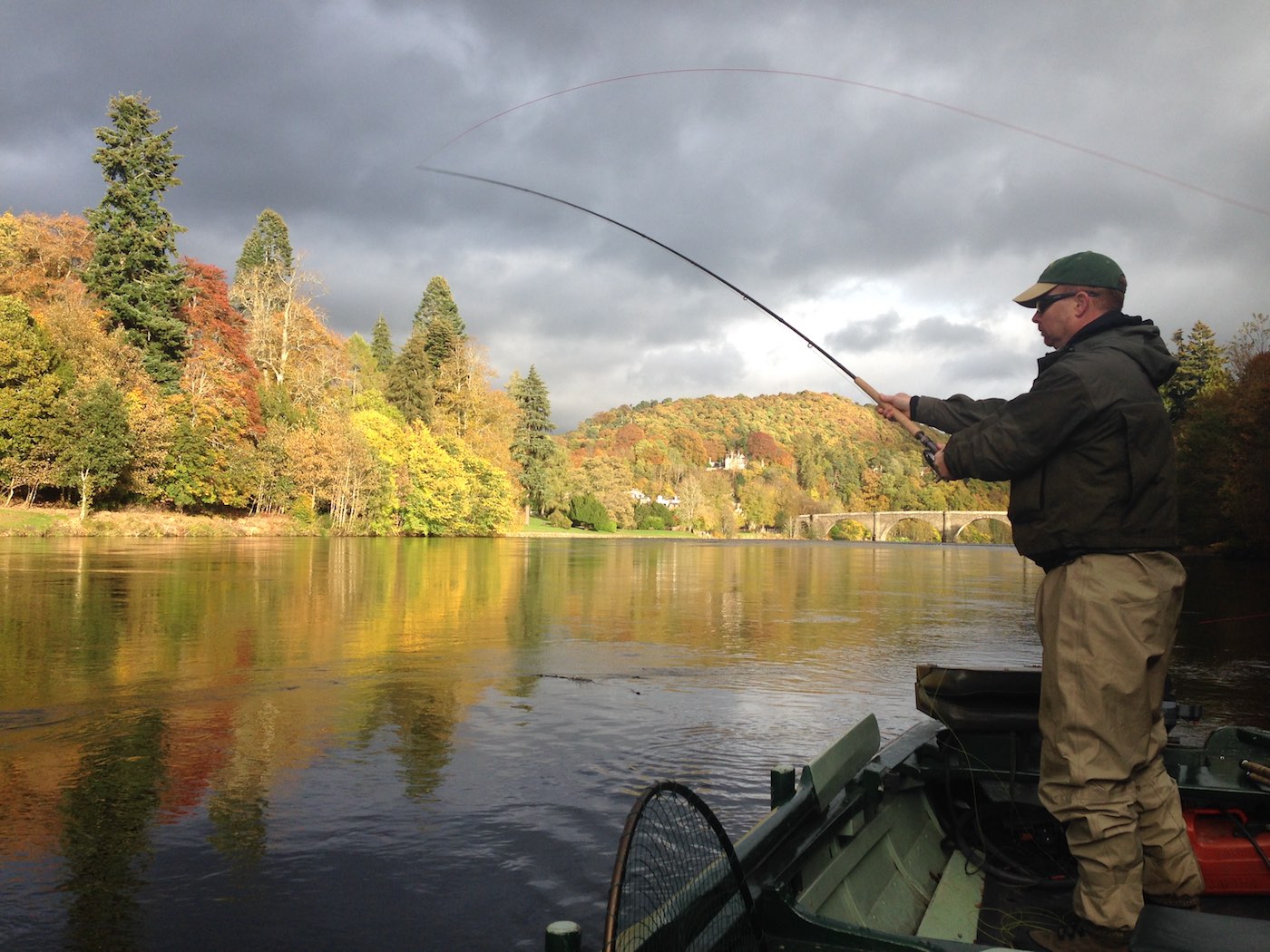 Here's a salmon fishing holiday guest playing a salmon on the Dunkeld House beat of the River Tay. This is a traditional salmon fishing boat the fishing trip guest is using as a casting platform and to get him closer to the long distance salmon lies. You can see the sandstone bridge in Dunkeld in the distance being illuminated by the late afternoon sunlight.
