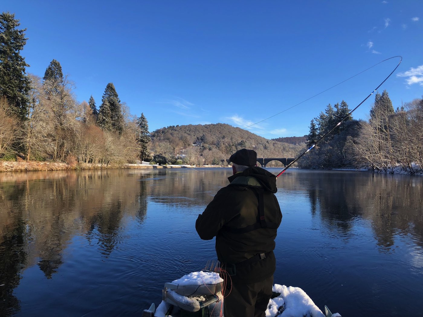 This River Tay salmon trip guest is fishing from the traditional Tay salmon fishing boat during late January on the Tay at Dunkeld. If you look closely you'll see the 200 year old Telford Bridge in the distance which is one of the Tay's most historic & beautiful sandstone bridges. This fine shot was taken by the fisher's personal Tay salmon guide and boatman for the day.