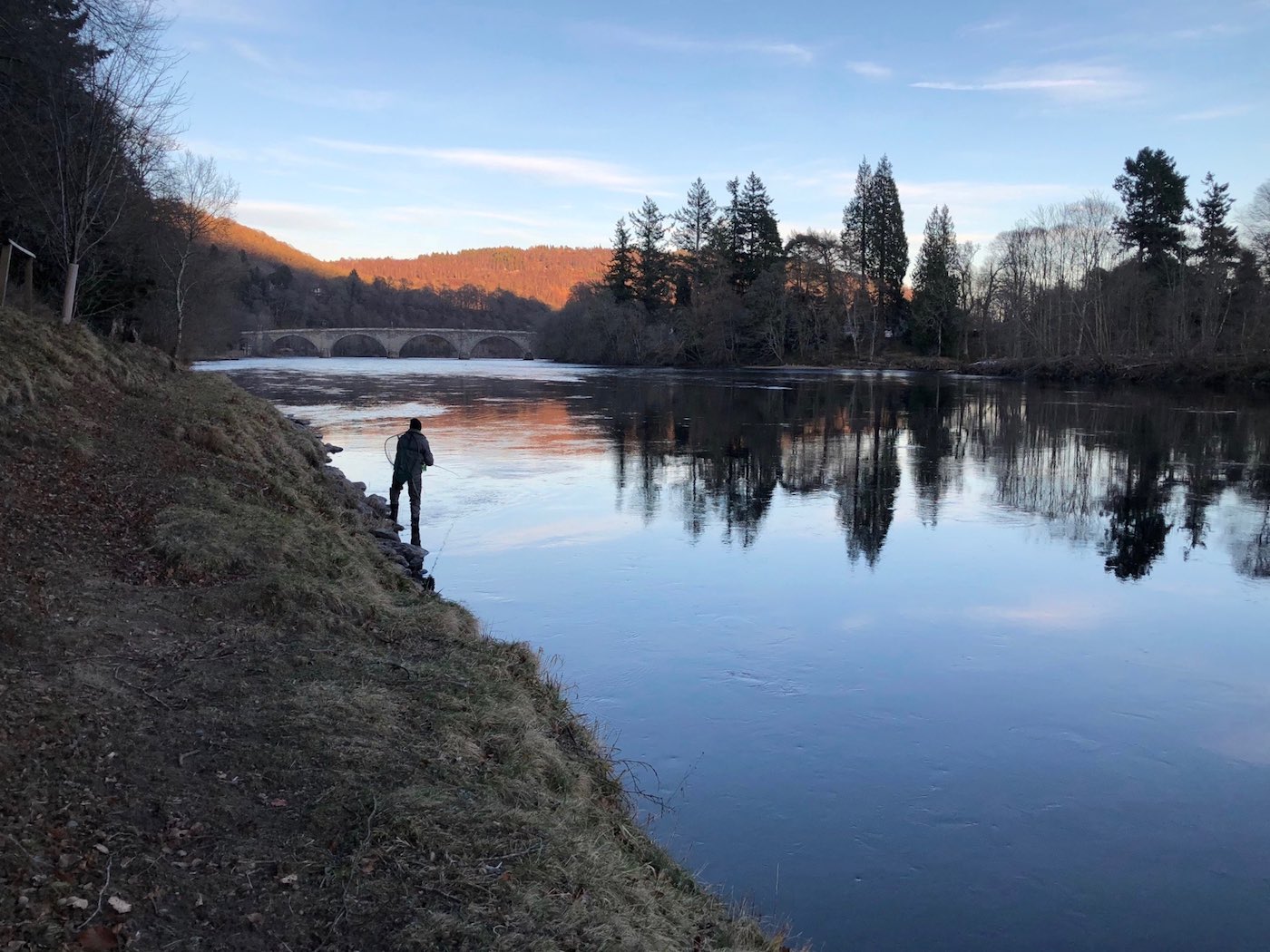 This is the tail of the Gauge Tree Pool on the Dunkeld House salmon fishing beat of the River Tay slightly upstream of the historic Dunkeld Bridge which you can clearly see in this photograph. I took this shot of one of our River Tay salmon fishing trip guests while working as his salmon fishing guide on the Tay. This shot was taken at dusk and you can see the sunset still illuminating the hills behind the bridge.