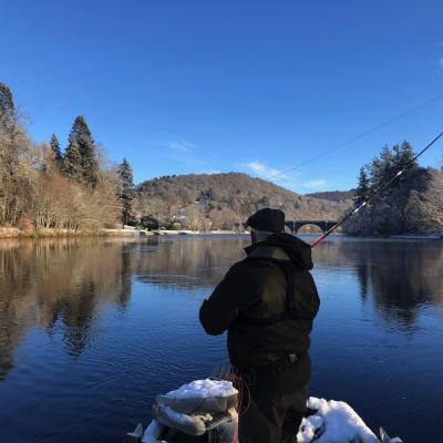 This River Tay salmon trip guest is fishing from the traditional Tay salmon fishing boat during late January on the Tay at Dunkeld. If you look closely you'll see the 200 year old Telford Bridge in the distance which is one of the Tay's most historic & beautiful sandstone bridges. This fine shot was taken by the fisher's personal Tay salmon guide and boatman for the day.