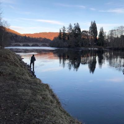 This is the tail of the Gauge Tree Pool on the Dunkeld House salmon fishing beat of the River Tay slightly upstream of the historic Dunkeld Bridge which you can clearly see in this photograph. I took this shot of one of our River Tay salmon fishing trip guests while working as his salmon fishing guide on the Tay. This shot was taken at dusk and you can see the sunset still illuminating the hills behind the bridge.